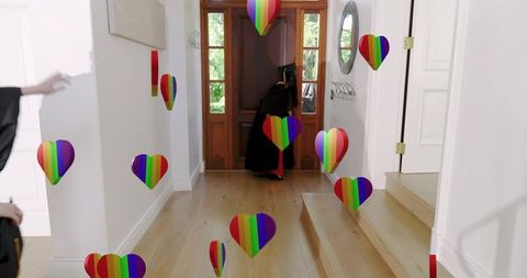 Walking graduate wearing cap and gown celebrating pride with rainbow heart confetti in home foyer