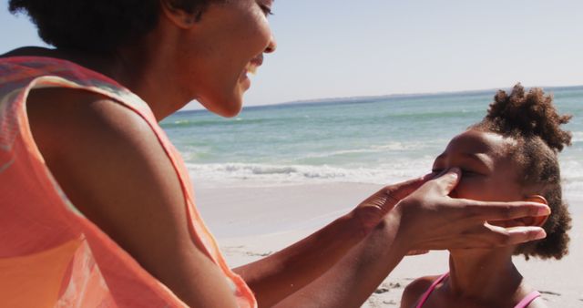 A mother applying sunscreen to her daughter while at the beach on a sunny day. Smiling and enjoying their time by the ocean, this image captures a loving family moment focusing on skin protection and care during outdoor activities. Ideal for use in articles on family vacations, skin care tips, parental guidance, and summertime activities.