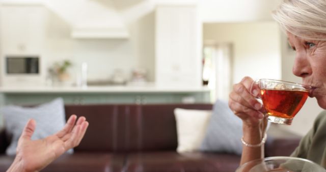 Senior woman drinking tea while sitting on a couch in the living room, chatting with a friend. Perfect for themes involving relaxation, friendship, home lifestyle, and senior living.