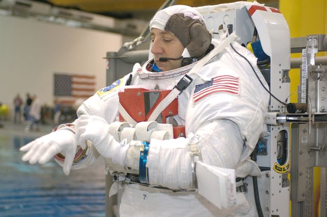 An astronaut is preparing for extravehicular activity (EVA) by putting on a training version of the Extravehicular Mobility Unit (EMU) spacesuit at the Neutral Buoyancy Laboratory near the Johnson Space Center. The image evokes themes of space exploration, astronaut training, and scientific study, making it suitable for educational material, space agency publications, scientific articles, and content related to space missions and astronaut training programs.
