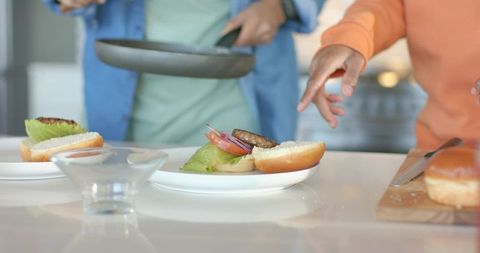 Friends assembling juicy burgers on glossy kitchen counter, cooking together at home