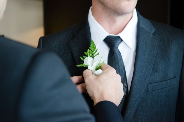 Man in formal suit receiving boutonniere adjustment before wedding ceremony, symbolizing love and commitment. Ideal for use in wedding blogs, preparation guides, and event planning materials.
