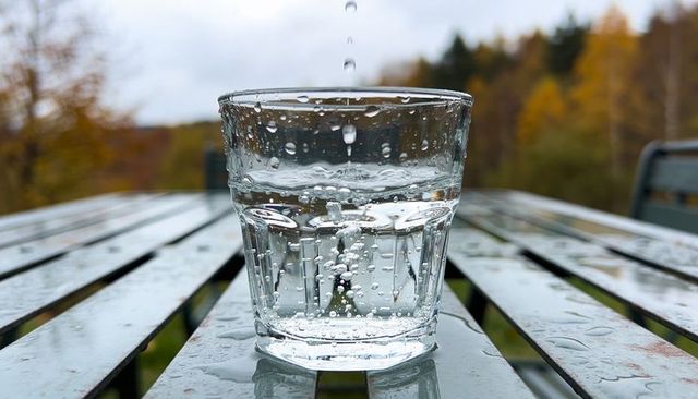 Raindrops splashing into glass of water on wet patio table with autumn trees