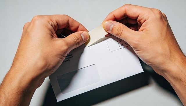 Male hands peeling open white window envelope on gray desk, revealing mail document