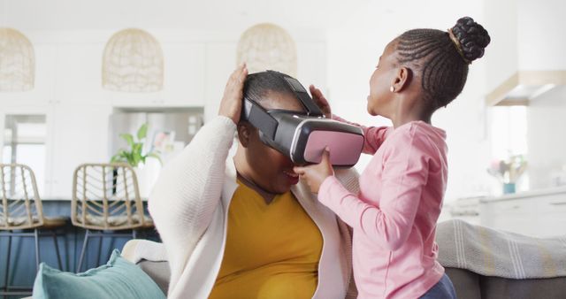 An African American mother and her daughter enjoying a fun moment exploring virtual reality technology together. Ideal for use in ads or content promoting family activities, technology use at home, parenting, and modern living spaces.