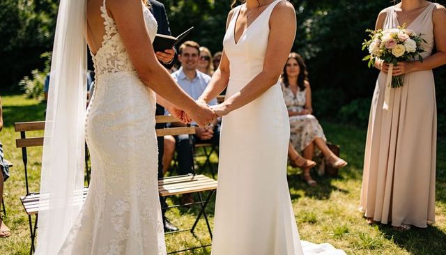 Two brides holding hands during outdoor garden vow exchange, elegant lace and modern gown
