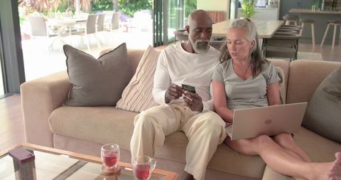 Mature mixed-race couple sitting on sofa checking credit card and laptop in living room