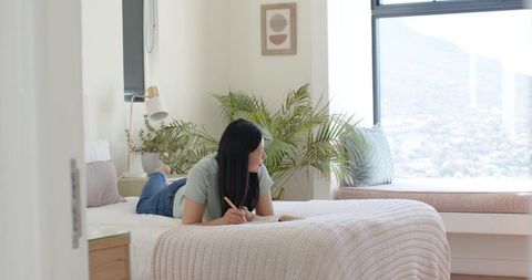 Asian woman lying on bed writing in notebook by window in cozy minimalist bedroom