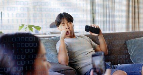 Yawning man holding smartphone on sofa with binary code overlay, partner filming casual home