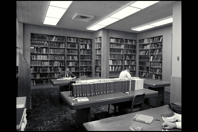 Classic black and white scene inside a vintage library with people reading and working at tables. Shelves filled with books surround the room, creating a cozy and intellectual atmosphere. Ideal for illustrating themes of academia, research, history, and the timeless value of libraries. Useful for educational resources, historical articles, and literature blogs.
