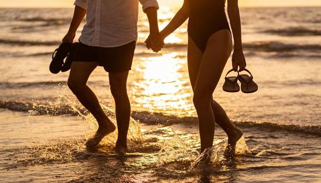 Couple walking hand-in-hand through ocean waves at golden hour holding flip-flops