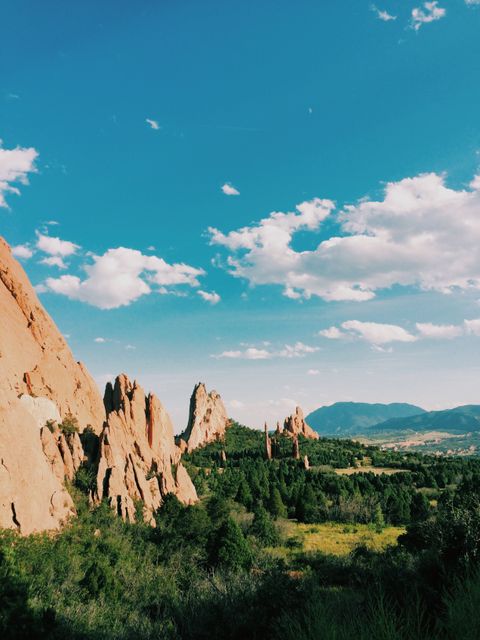 Stunning rock formations basking in sunlight with vibrant greenery and a bright blue sky with scattered clouds. Ideal for use in travel brochures, tourism websites, nature magazines, and outdoor adventure marketing.