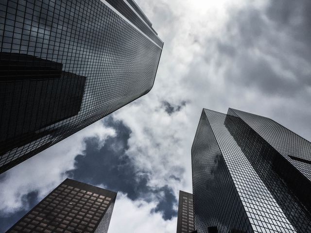 This image features a dramatic low angle view of modern skyscrapers reaching towards a cloudy sky. The glass facades of the buildings reflect the atmospheric clouds creating an urban and modern vibe. Suitable for use in business advertisements, architecture blogs, urban planning presentations, and any media related to business districts and modern city living.