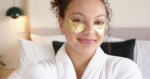 Smiling african american woman wearing gold eye patches white robe skincare ritual