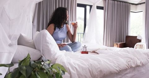 African american woman sitting on canopy bed holding teacup in bright cozy bedroom