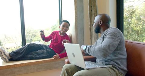 African american father and son bonding and chatting by window with laptop and smartphone