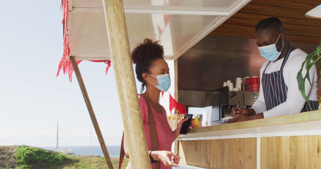Outdoor street food stall showing a female customer in a pink top interacting with a male vendor wearing striped apron and face mask. Useful for illustrating pandemic safety measures, street food culture, or outdoor dining scenarios during COVID-19.