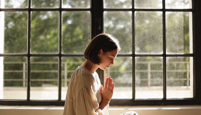 Young woman praying and meditating by sunlit grid window, soft backlight, serene moment