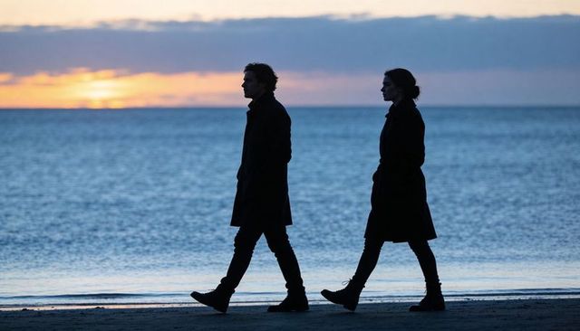Silhouetted couple walking twilight beach in long coats and boots during ocean sunset