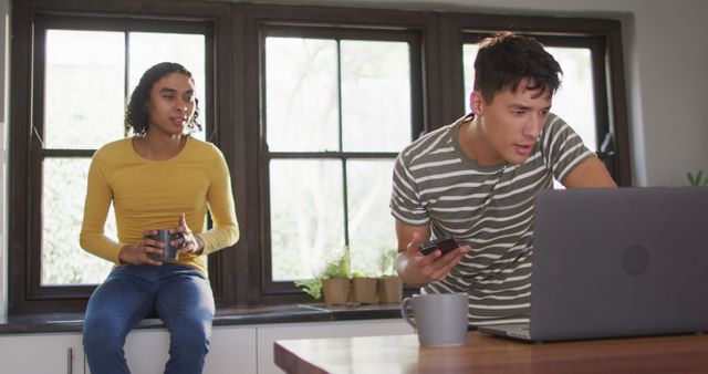 This image shows two young adults in a home office setting. One individual is working on a laptop while multitasking with a phone, suggesting focus and efficiency. The other is holding a coffee mug, indicating a relaxed discussion in a modern work environment. Ideal for illustrating concepts of remote work, young professionals, collaboration, and a casual work atmosphere.