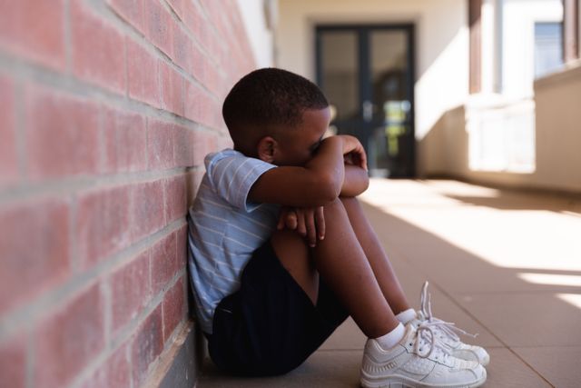 Side view of african american elementary schoolboy hugging knees while sitting on floor in corridor. unaltered, education, sadness, depression, failure, loneliness and school concept.