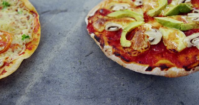 Two homemade veggie pizzas with fresh toppings are placed on a stone surface. The pizzas are adorned with slices of avocado, mushrooms, tomatoes, and cheese. Perfect image for use in advertisements, food blogs, restaurant menus, or healthy meal campaigns.