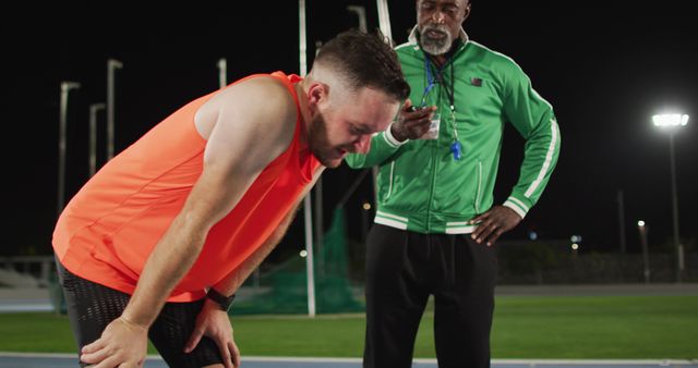 Man wears bright athletic clothes, bent over from physical exertion, indicating intense training. Coach stands nearby wearing green jacket, looking supportive and motivating, in an outdoor sports facility lit at night. Perfect for illustrating themes of hard work, endurance, athletic training, motivation, and coach-athlete relationships.