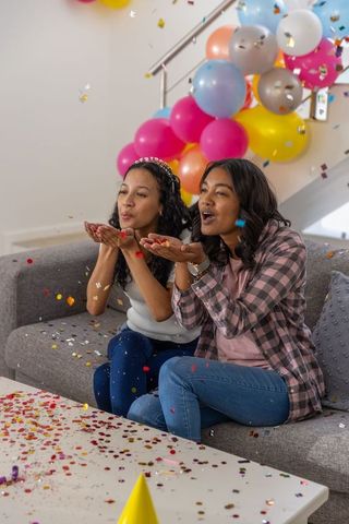 Friends blowing metallic confetti while celebrating birthday with colorful balloons indoors