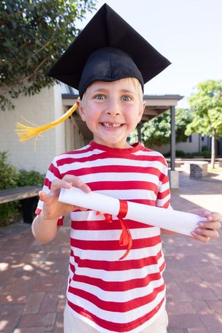 Smiling boy celebrating kindergarten graduation wearing mortarboard holding ribbon-tied diploma