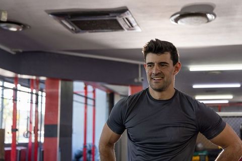 Mid-adult man standing in gym wearing dark grey t-shirt hands on hips confident expression