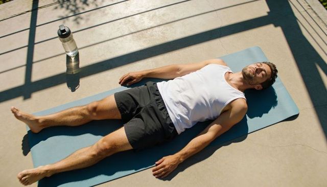 Young man resting on yoga mat on sunlit balcony, meditating and hydrating after workout