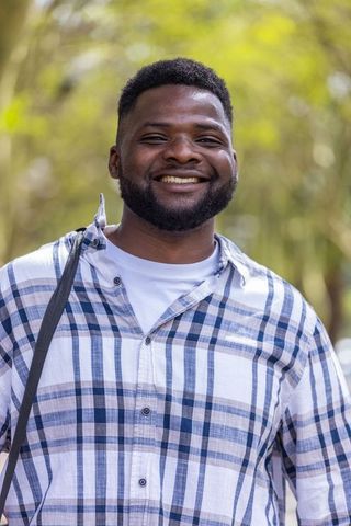 African man smiling standing in park wearing plaid shirt and shoulder strap, casual lifestyle