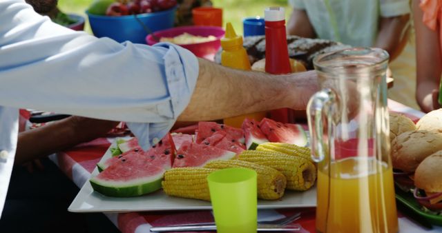 A diverse group of people is gathered around a picnic table filled with summer treats like watermelon and corn, with copy space. It's a casual outdoor setting that suggests a friendly gathering or family barbecue.