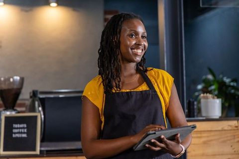 Smiling barista holding tablet behind espresso bar, serving coffee in warm industrial cafe