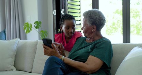 African grandmother and granddaughter sharing mobile phone on sofa teaching and bonding