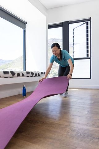 Asian woman unrolling purple yoga mat in bright sunlit home practicing yoga and stretching