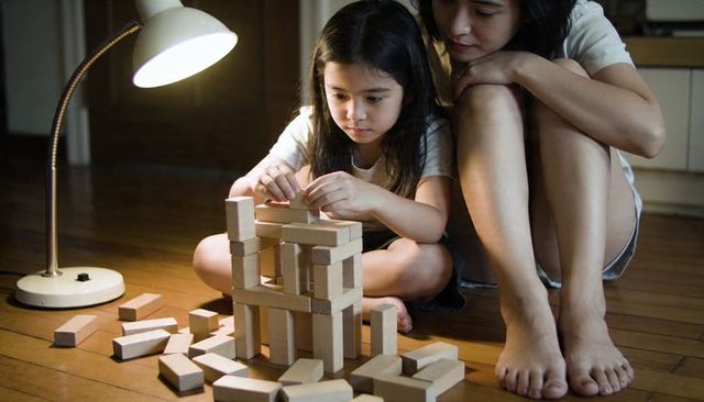 Mother and child building wooden block tower on hardwood floor under lamp glow