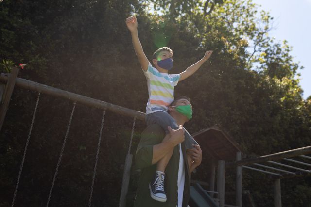 Side view of a caucasian man carrying his son on his shoulders at a park while his son is raising his hands up high on a bright and sunny day. behind them are swings and monkey bars.