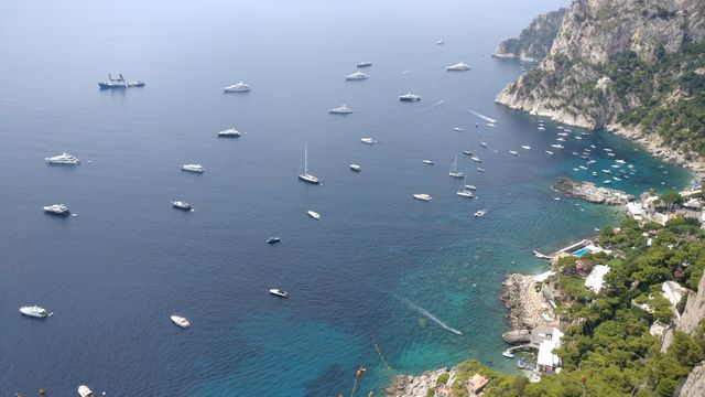 Numerous boats and yachts seen from above on clear blue water near a rocky coastline. Ideal for travel brochures, Mediterranean tourism promotions, scenic wallpapers, vacation advertisements, and nautical themed posters.