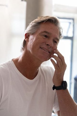 Senior man touching cheek and smiling by window wearing white t-shirt and smartwatch, natural light