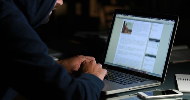 Hooded figure typing on modern laptop in dimly lit environment, suggesting secretive activities. Glowing screen illuminates keyboard, emphasizing reliance on technology. Image can be utilized in articles about cybersecurity, cybercrime, and data protection, or in presentations focusing on internet safety and programming.