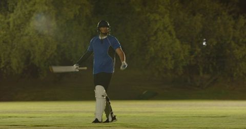 Night cricket batsman walking off after dismissal under floodlights on grassy pitch