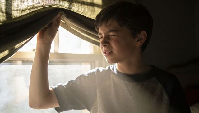 Teenage boy squinting while lifting curtain, warm sunbeam through window