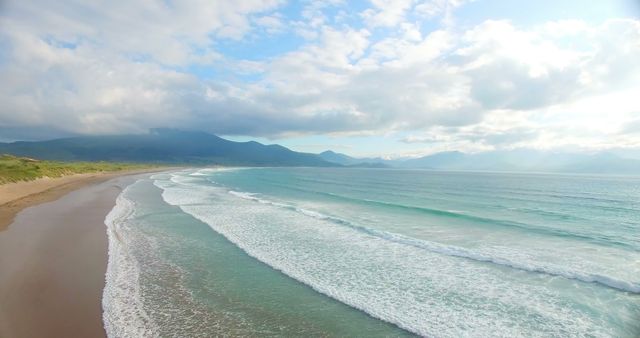 A serene beach landscape showcases the gentle waves of a turquoise sea meeting a sandy shore, with mountains in the distance under a vast sky. The tranquility of the scene invites viewers to imagine the sound of the waves and the feel of the ocean breeze.