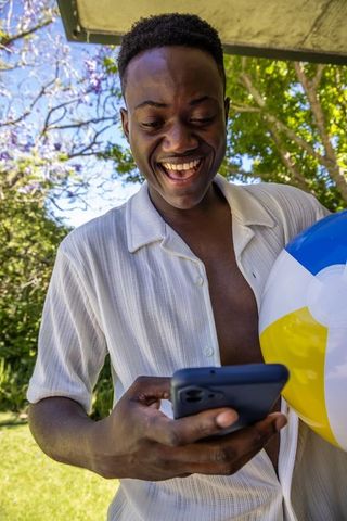 Joyful african man checking smartphone on porch holding beach ball in sunny backyard