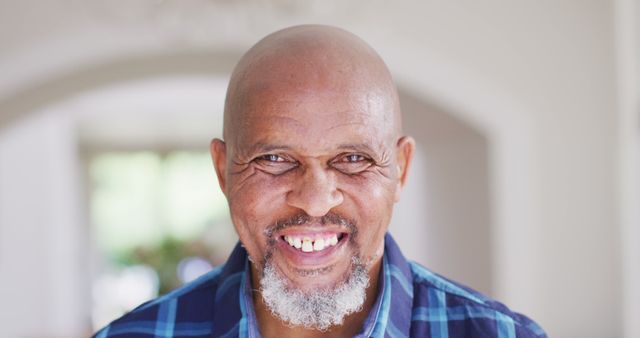 Capturing an older African American man with a big smile, wearing a blue plaid shirt. His bald head and friendly expression convey warmth and positivity. The indoor setting with a blurred background makes this usable for content on seniors' wellbeing, positivity in aging, or general lifestyle topics focused on happiness and comfort.