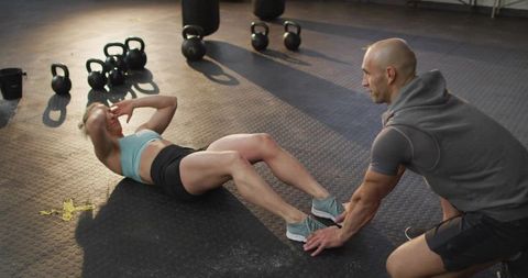Coach Holding Feet While Woman Doing Sit-Ups in Gym with Kettlebells and Intense Lighting
