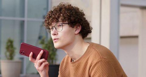 Curly-haired young man speaking into smartphone in urban courtyard wearing quilted sweater