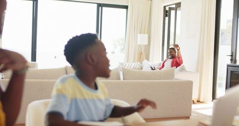 African american family relaxing at home with father waving from sofa and child reaching
