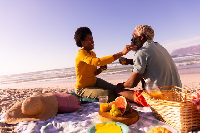 Happy african american mature woman feeding to senior man at beach against clear sky, copy space. nature, food, unaltered, love, togetherness, retirement, picnic, enjoyment and holiday concept.